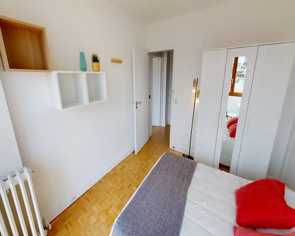 Chambre compacte et lumineuse avec lit double partiellement visible, armoire blanche à porte miroir, parquet et étagères décoratives minimalistes.