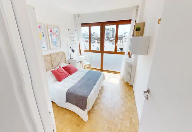 Chambre lumineuse et moderne avec grandes fenêtres, lit double bien fait avec coussins décoratifs, petit bureau près de la fenêtre et parquet chaleureux — idéale pour un séjour citadin confortable.