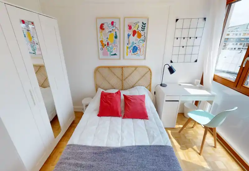 Chambre lumineuse et bien aménagée avec lit complet, deux coussins rouges, tête de lit en rotin et bureau ensoleillé près de la fenêtre — parfaite pour les locations courte durée.