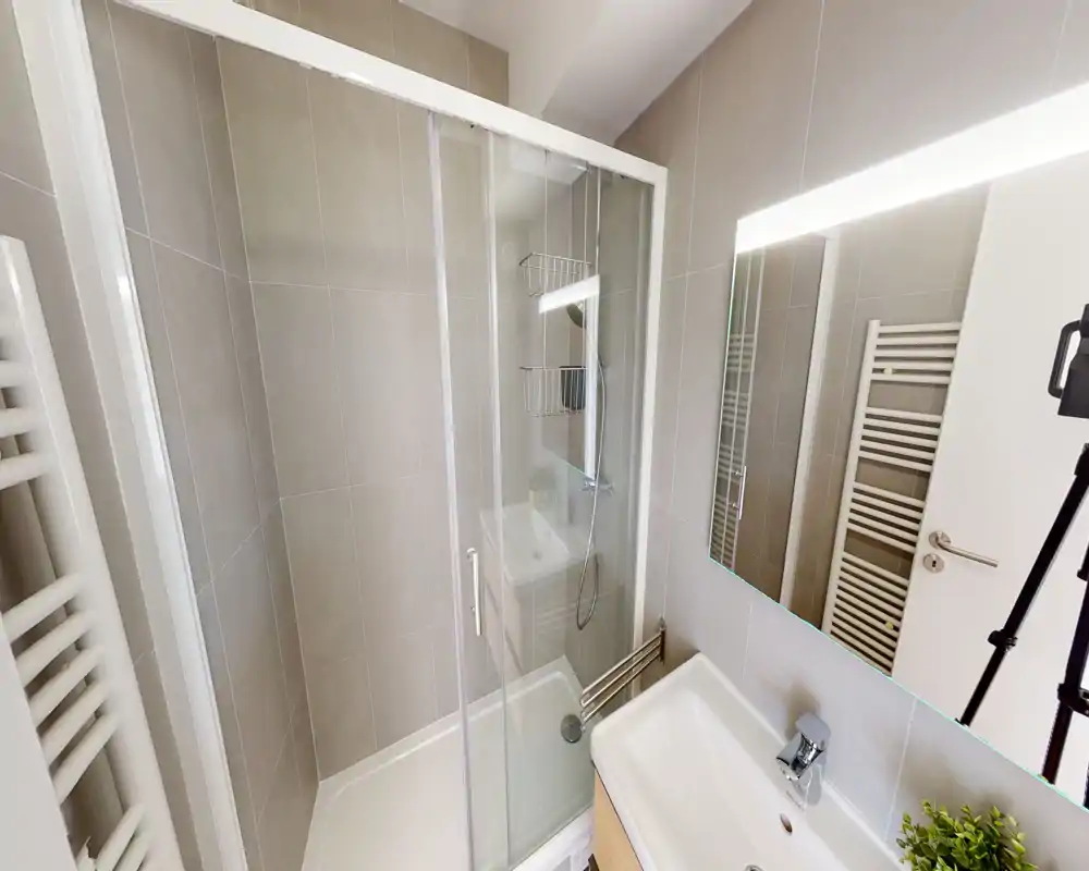 Salle de bain moderne et compacte avec douche vitrée, lavabo sur meuble et miroir éclairé ; carreaux gris neutres et radiateur sèche-serviettes pour un rendu propre et contemporain.