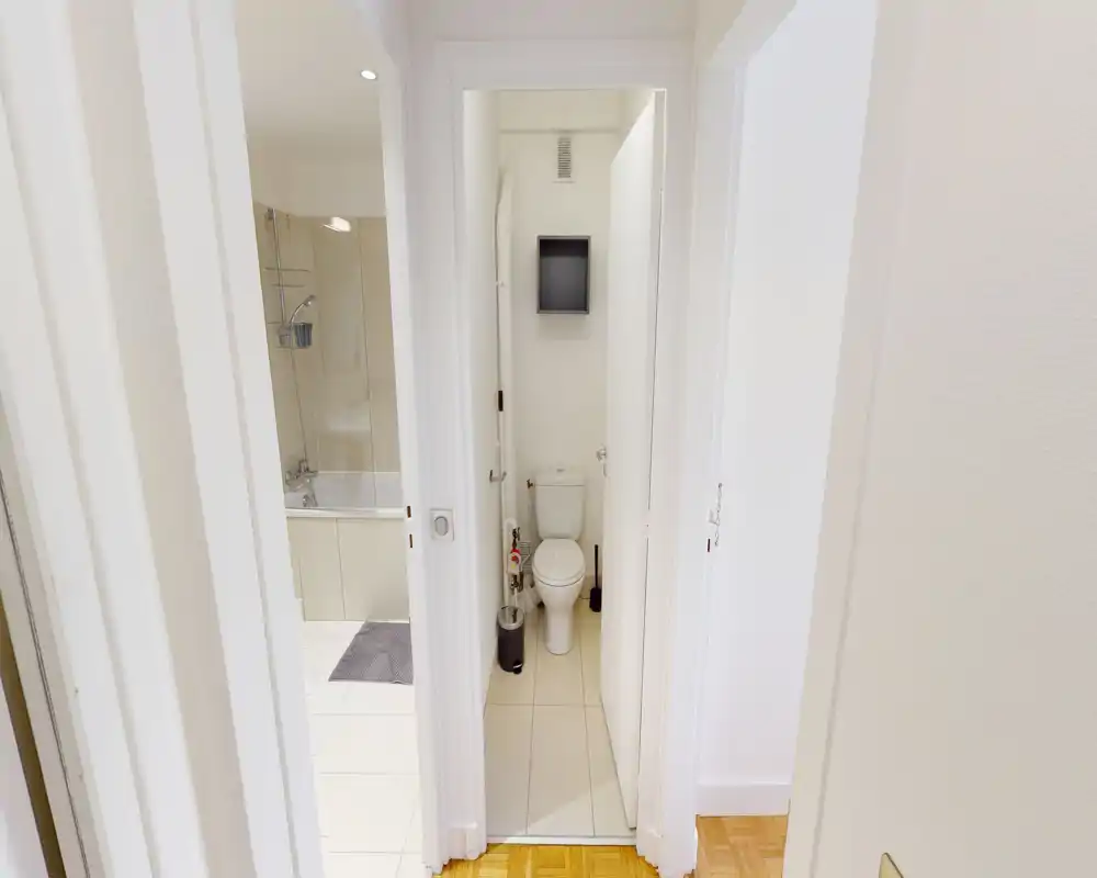 Salle de bains compacte et propre avec des toilettes dans un WC étroit séparé et un aperçu d'une baignoire/douche adjacente ; finitions blanches et sol carrelé.