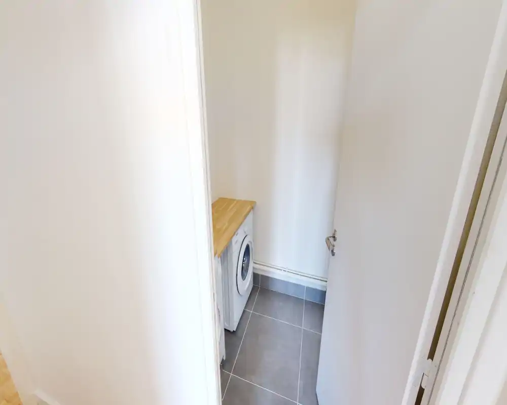 Petit coin buanderie/utility avec lave-linge frontal sous un plan de travail en bois ; sol carrelé et murs blancs pour un espace propre et fonctionnel.