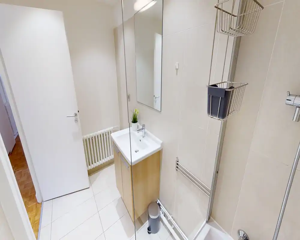 Salle de bains compacte et moderne avec lavabo mural, miroir, zone de douche et carrelage beige neutre. Espace lumineux et propre, idéal pour les locations courtes durées.