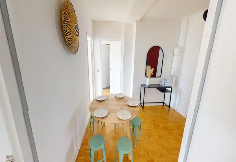Coin repas moderne et lumineux avec une table ronde en bois dressée pour quatre et des tabourets vert menthe, accompagné d'une console et d'un miroir décoratif — un espace compact et accueillant.
