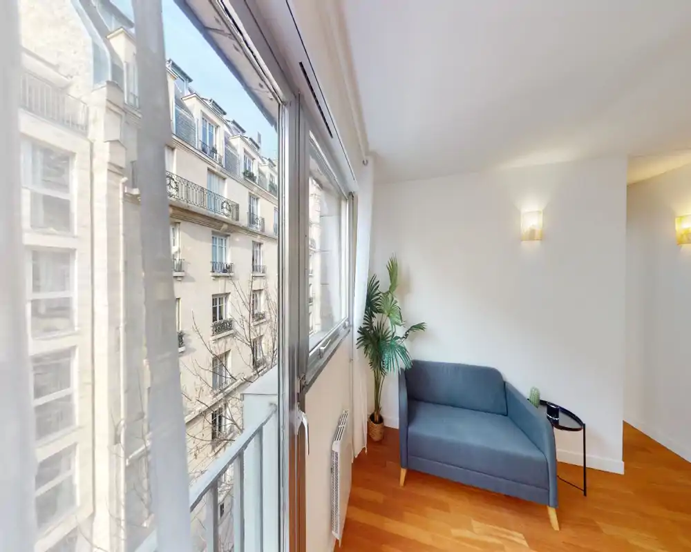 Espace de vie lumineux et moderne avec un petit canapé bleu, une table d'appoint et une grande plante près d'une large fenêtre donnant sur des immeubles — idéal pour un studio chaleureux.