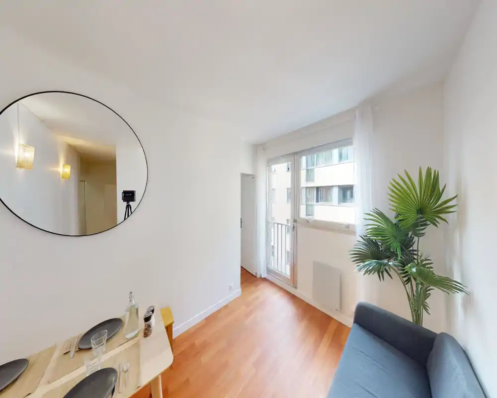 Espace de vie lumineux et minimaliste avec un petit canapé, une plante décorative et un coin repas près d’une grande fenêtre apportant beaucoup de lumière naturelle.