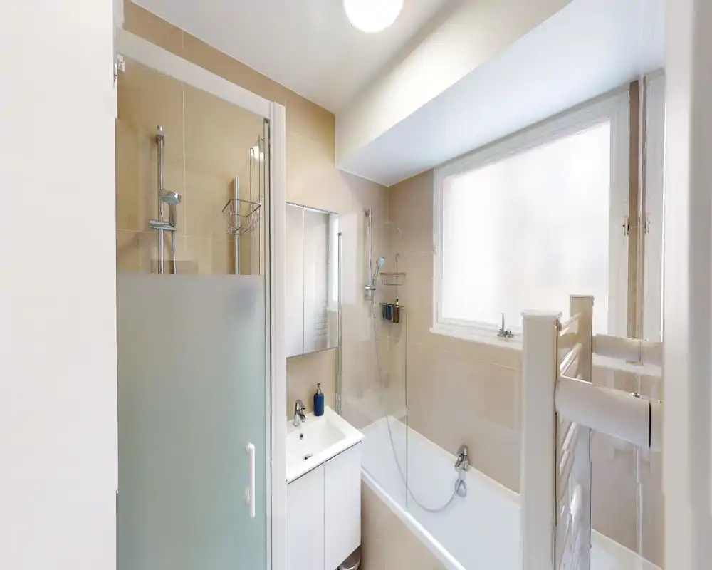 Salle de bains moderne et lumineuse avec baignoire, douche, porte de douche en verre dépoli et meuble-vasque. Carrelage beige neutre et grande fenêtre dépoli offrant intimité et lumière douce.