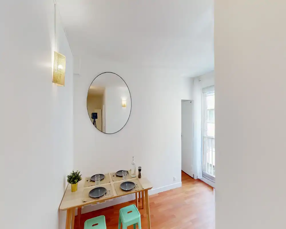Coin repas lumineux et compact avec une table en bois dressée pour quatre, un grand miroir rond et la lumière naturelle d'une fenêtre — style moderne et épuré adapté aux petits appartements.