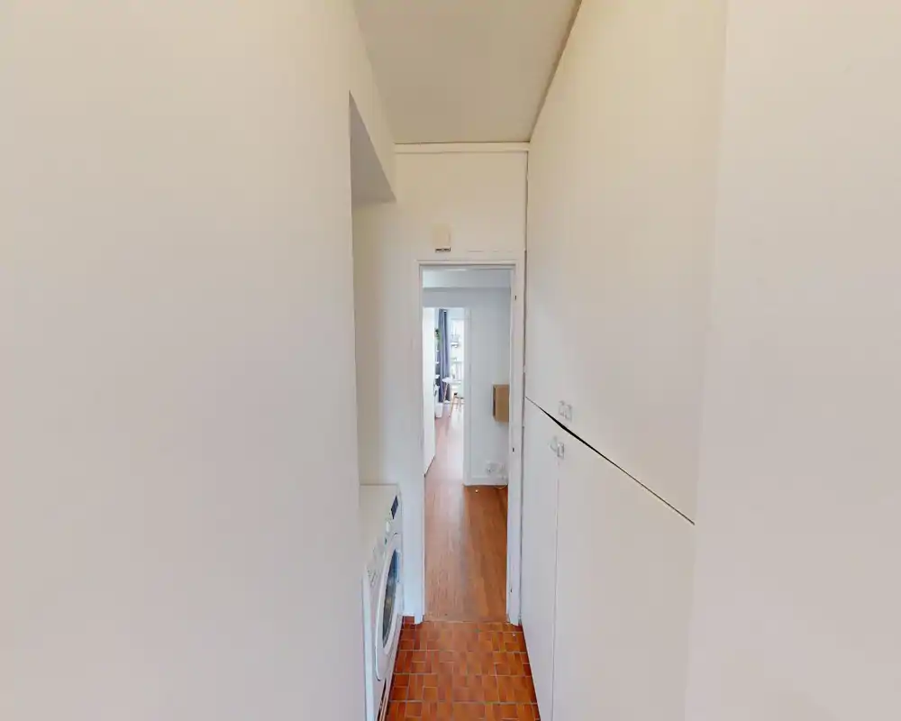 Entrée/couloir étroit avec rangements intégrés et machine à laver, menant au séjour. Couloir lumineux et fonctionnel, adapté à un usage pratique.