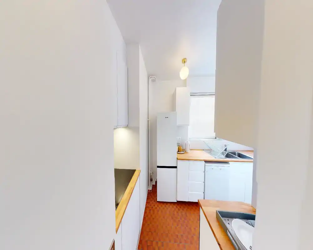 Cuisine compacte et lumineuse avec placards blancs, plans de travail en bois et sol carrelé ; réfrigérateur, évier et lave-vaisselle dans un agencement pratique.