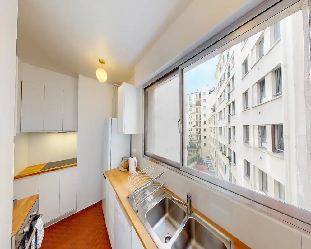 Cuisine en enfilade compacte et lumineuse avec plans de travail en bois, évier inox et grande fenêtre donnant sur les immeubles voisins. Placards blancs et éclairage chaleureux offrent un espace propre et fonctionnel.