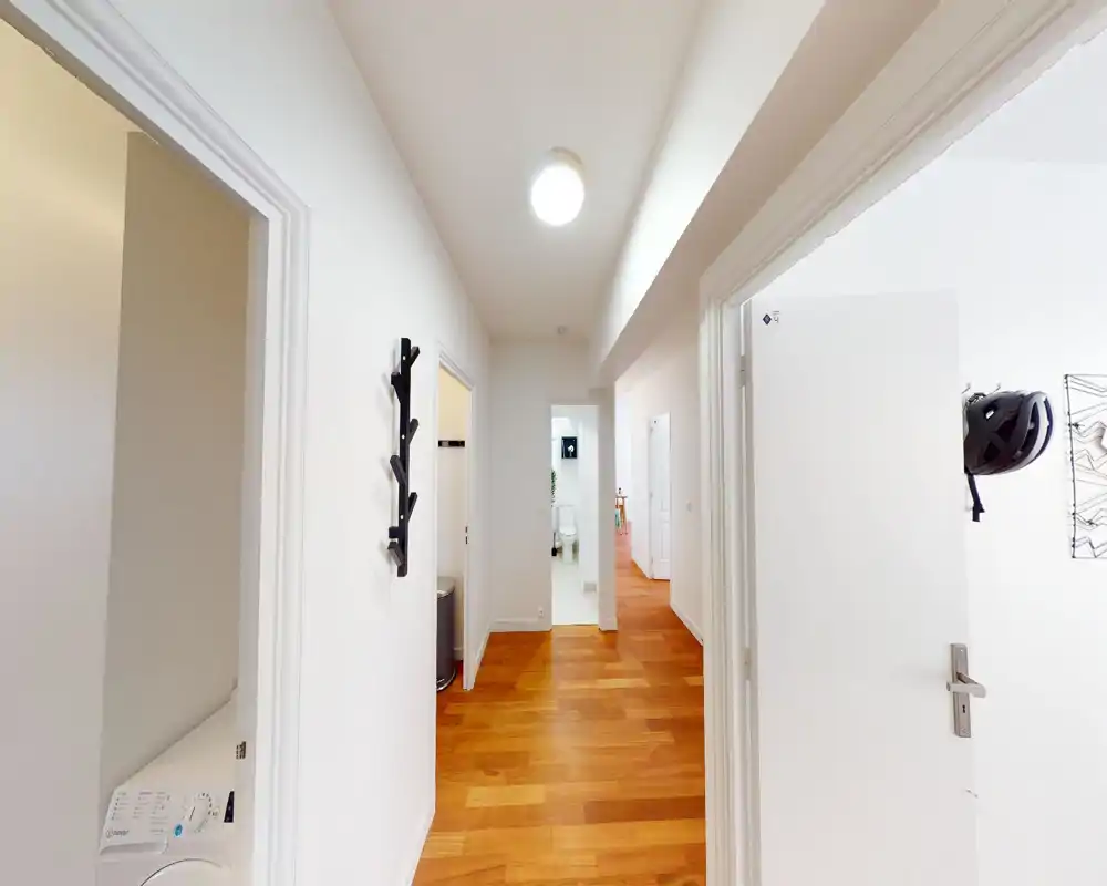Entrée lumineuse et propre avec un sol en bois chaleureux, des murs blancs et plusieurs portes donnant sur d'autres pièces. Un porte-manteau mural et une lampe au plafond créent un espace accueillant.