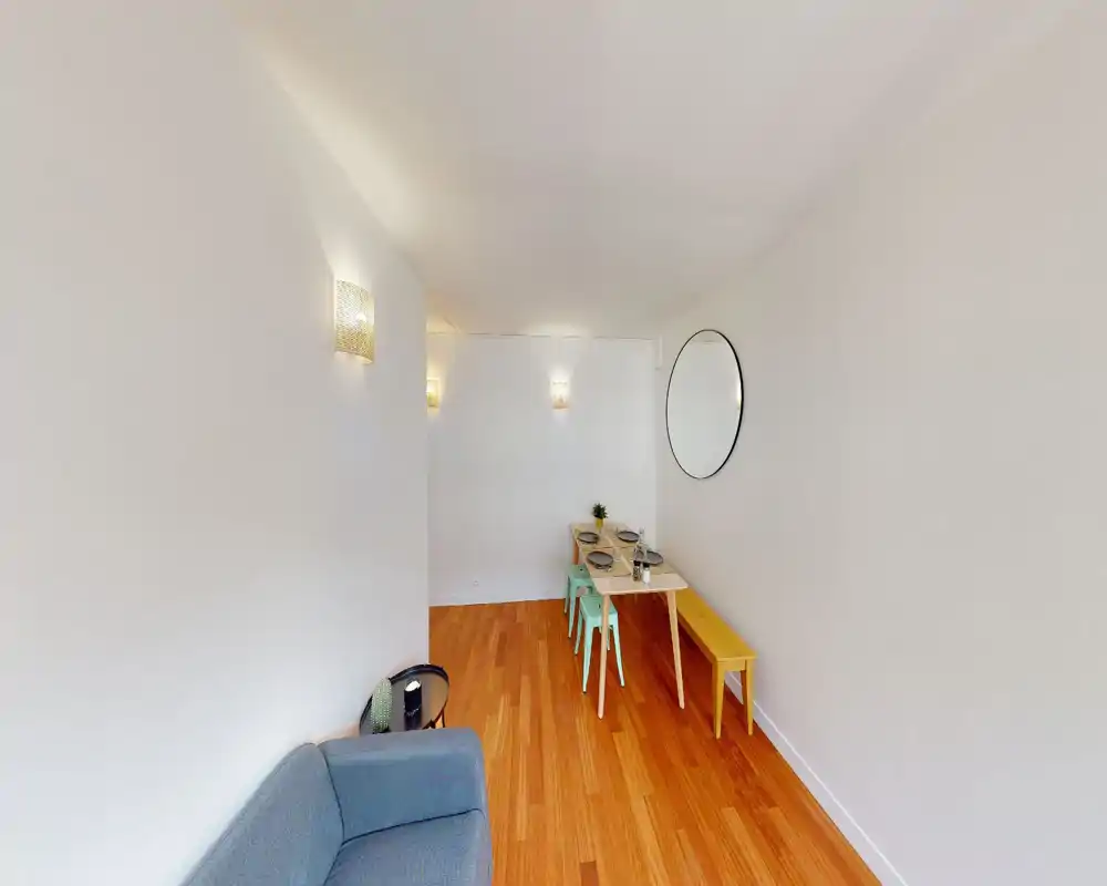 Espace jour lumineux et compact avec un petit canapé, une table à manger simple et un banc, un miroir rond décoratif et un sol bois chaleureux — idéal pour un appartement urbain cosy.