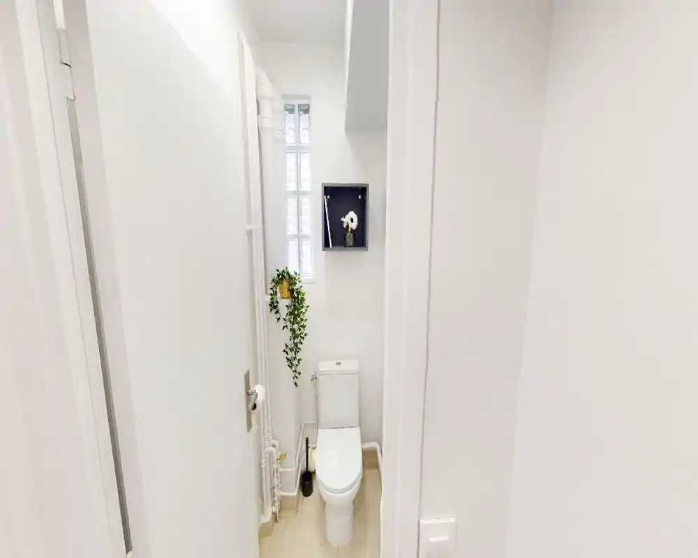Salle de bains compacte et lumineuse avec un WC blanc moderne, une petite fenêtre en pavé de verre, une étagère décorative et une plante pour un aspect frais et minimaliste.