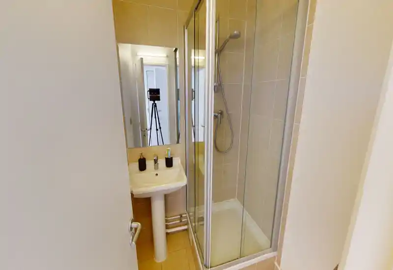 Compact tiled bathroom with a corner glass shower, pedestal sink and mirror above; bright, clean and efficiently arranged for a small space.