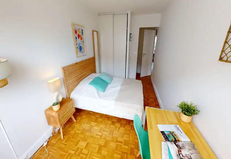 Bright, well-decorated bedroom with a wooden headboard, bedside table and a tidy desk. The room features parquet flooring, soft teal cushions and pleasant natural light — ideal for rental listings emphasizing comfort and style.