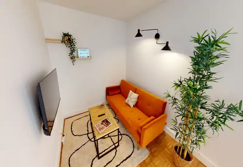 Cozy small living room with a bright orange sofa, wall-mounted TV, patterned rug and a tall potted plant — stylish and well-lit, ideal for relaxing.