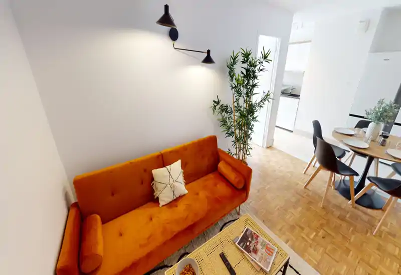 Espace de vie lumineux et élégant avec un canapé orange chaleureux, coussins décoratifs, une table basse et un petit coin repas visible vers la cuisine. Éclairage contemporain et plantes ajoutent une atmosphère cosy.