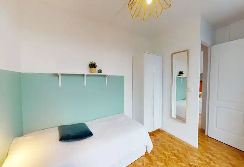Bright, compact bedroom with a single bed against a mint-green half-wall, parquet flooring, a tall white wardrobe and a full-length mirror — clean, minimal and welcoming.