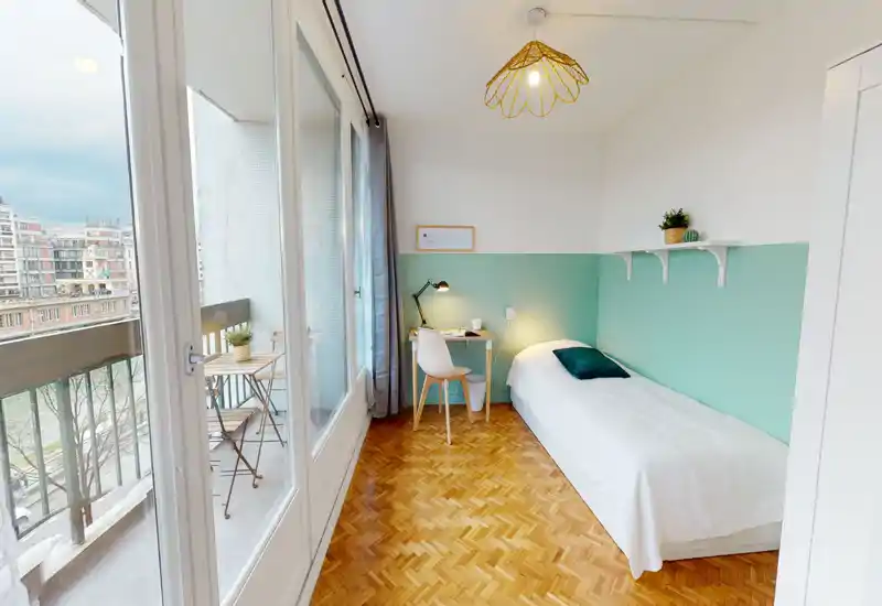 Bright, compact bedroom with a single bed, small desk and access to a balcony. Soft pastel wall, wooden parquet and ample natural light create a calm, welcoming atmosphere ideal for a single occupant or short stays.