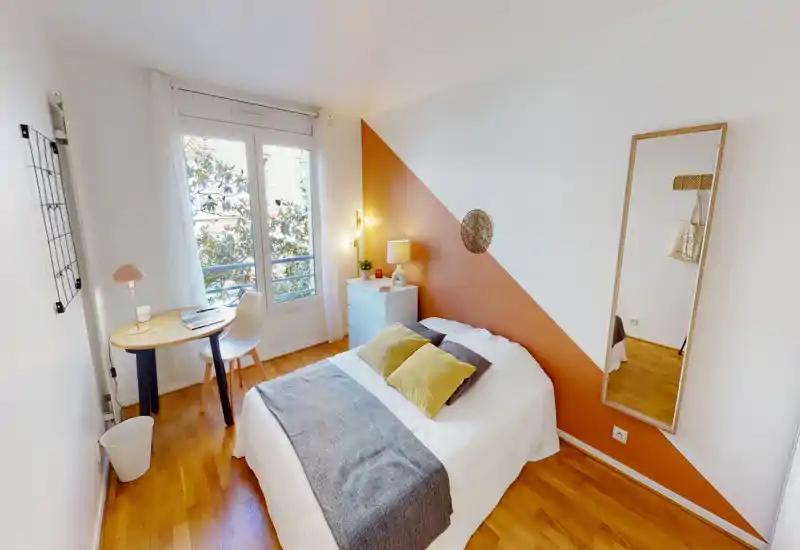 Chambre simple lumineuse et soignée avec un lit confortable, coussins décoratifs et un mur d'accent diagonal chaleureux. Grande fenêtre apportant lumière naturelle et petit bureau idéal pour se détendre ou travailler.