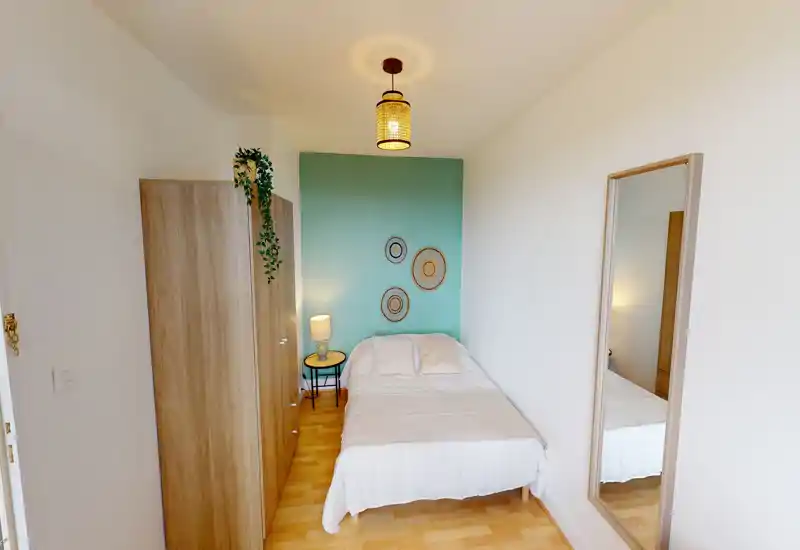 Chambre compacte et soignée avec un lit confortable devant un mur d'accent vert-bleu, armoire, table de chevet et grand miroir — lumineuse, bien décorée et idéale pour une annonce.