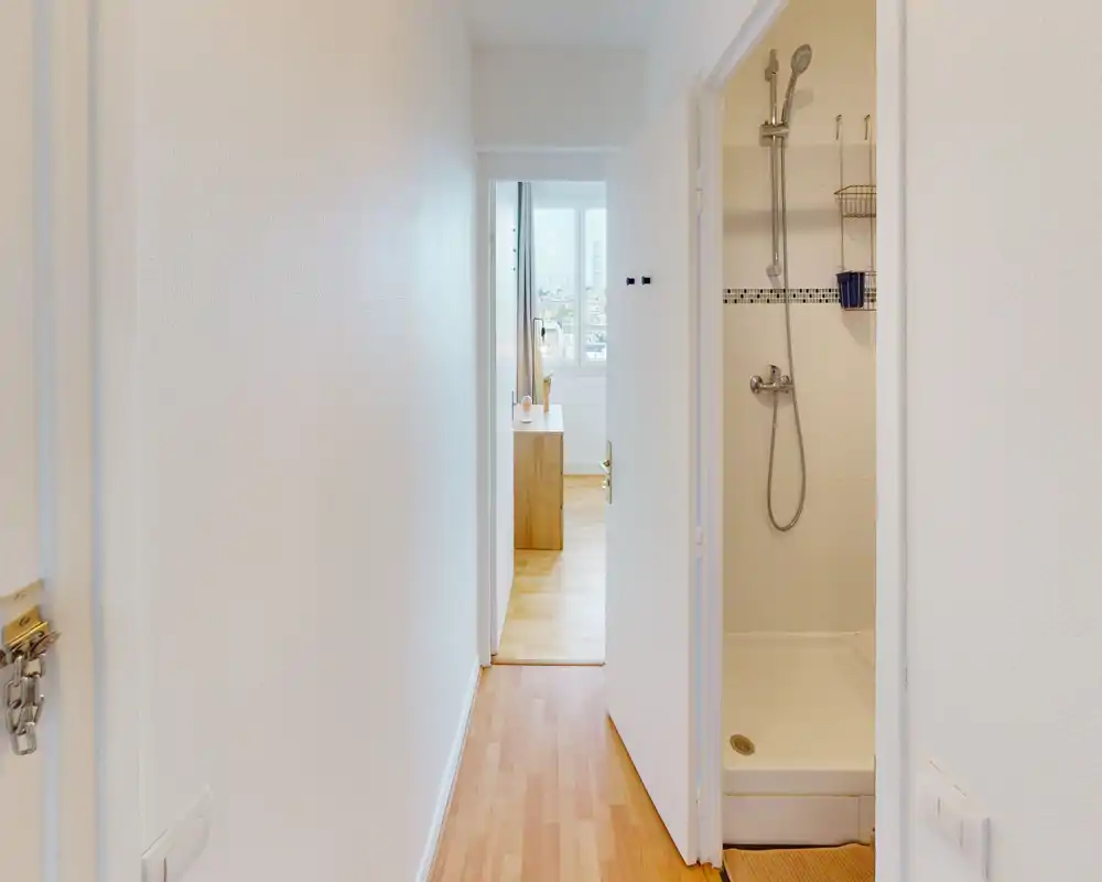 Couloir étroit et lumineux avec sol imitation bois menant à une pièce éclairée ; une salle de bains avec douche est visible sur la droite.