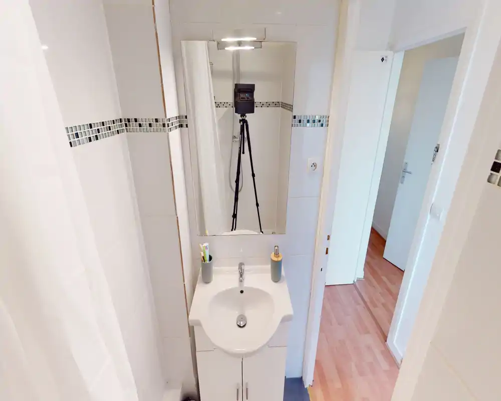 Salle de bain compacte et propre avec meuble-vasque blanc, miroir mural et rideau de douche ; carrelage blanc lumineux et fine frise en mosaïque.
