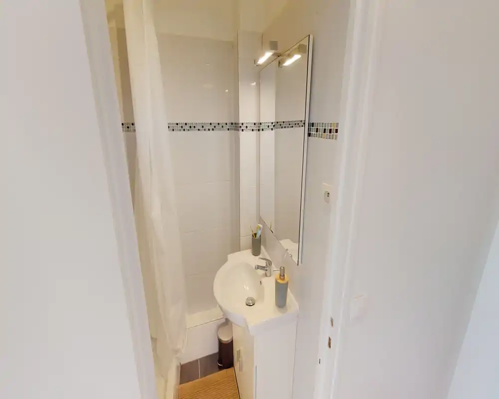 Salle de bains compacte et propre avec petit lavabo, miroir et douche avec rideau. Carrelage blanc et bandeau mosaïque apportent de la luminosité pour un espace fonctionnel.