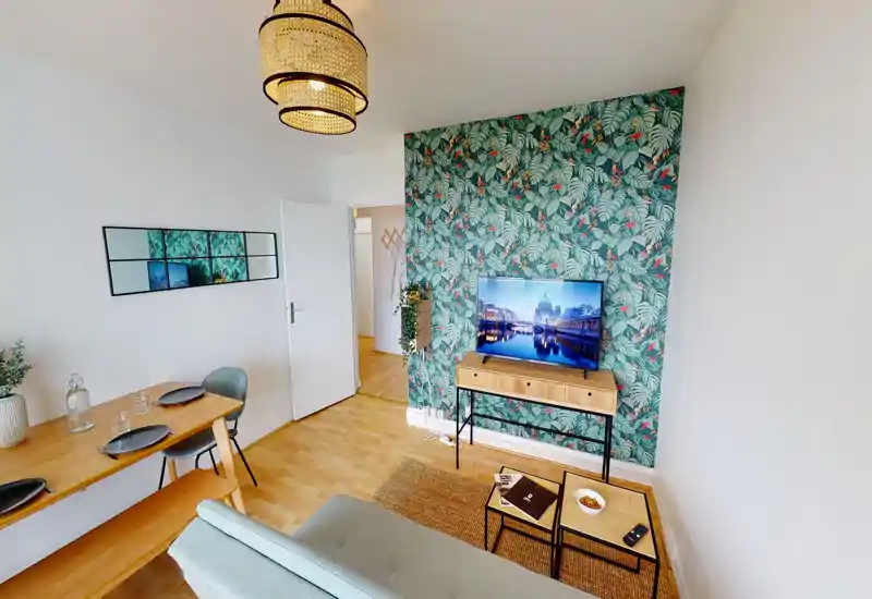 Séjour lumineux et soigné avec un mur d'accent à motif, petit canapé, TV sur console et coin repas compact — espace moderne et convivial, idéal pour un couple ou un voyageur seul.