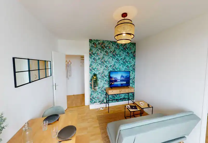 Salon lumineux et élégant avec mur à motifs, petit canapé, télévision sur console et coin repas — idéal pour un séjour urbain cosy.