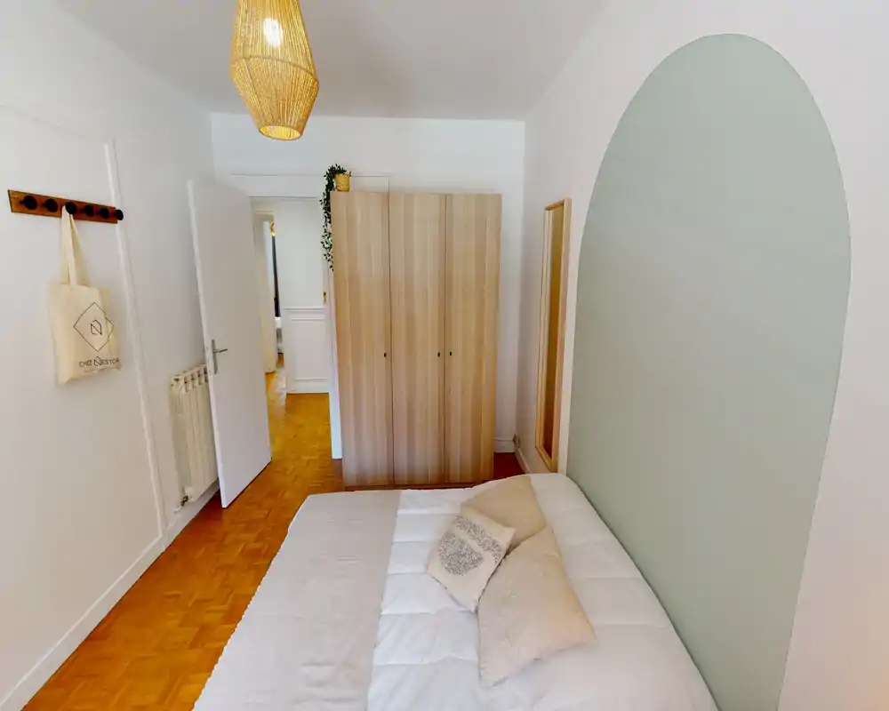 Chambre lumineuse et soignée avec lit double, coussins moelleux et armoire en bois ; une arche peinte pastel apporte du caractère et un luminaire suspendu crée une ambiance chaleureuse.