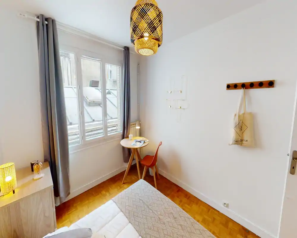 Bright, cozy bedroom with a neatly made bed, a small round desk by the window and warm decorative lighting — ideal for short stays or a single occupant.
