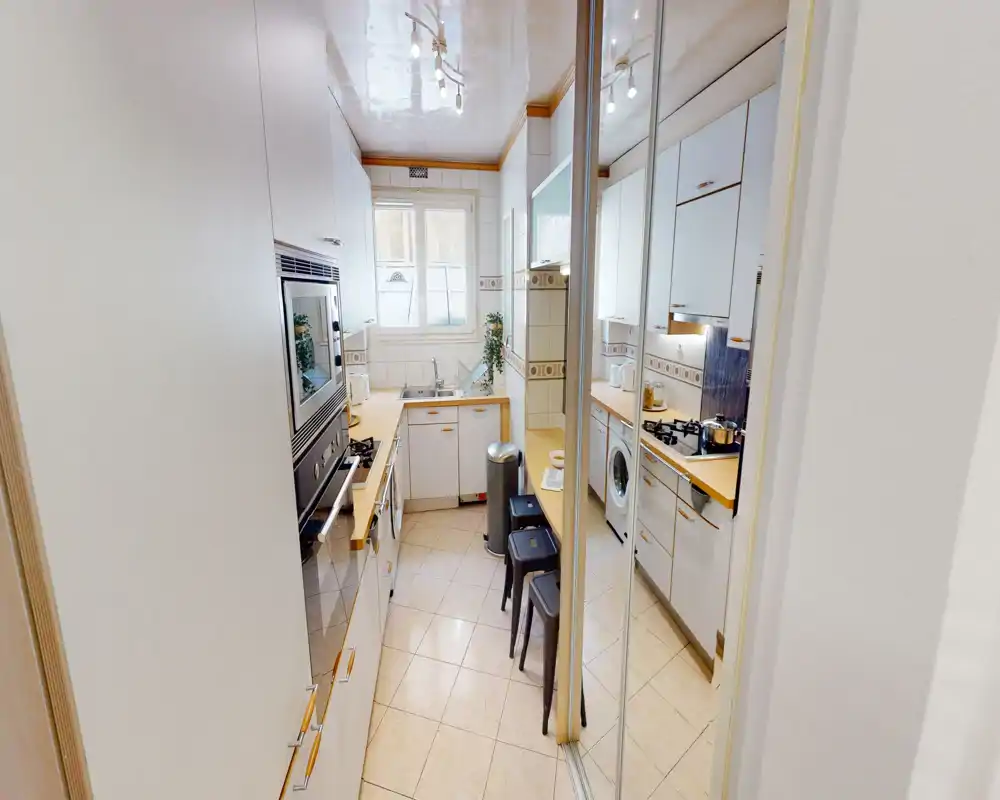 Compact, well-lit galley kitchen with white cabinets, wood-tone countertops, a gas hob and built-in appliances; mirrored doors and a window enhance the sense of space.