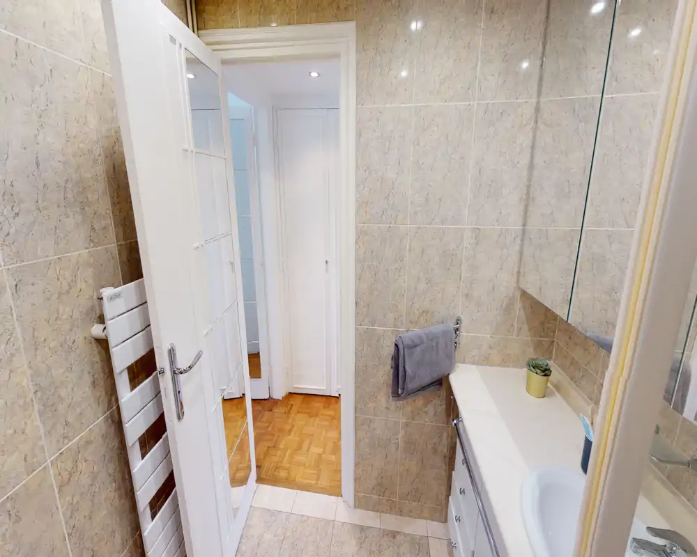 Bright, well-kept bathroom with marble-effect wall tiles, a sink countertop with storage, mirror cabinet and a heated towel rail by the door.