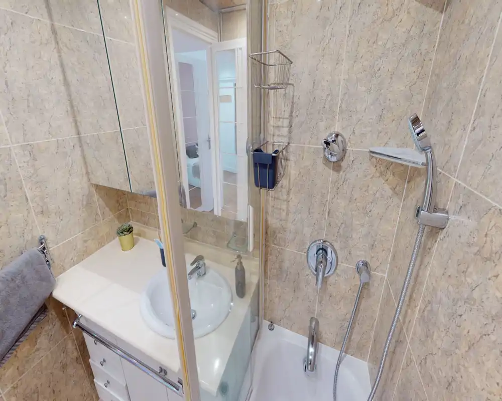 Compact, well-lit bathroom with a shower/bathtub combination, wall-mounted handheld shower, sink with vanity and tiled walls offering a clean, modern look.