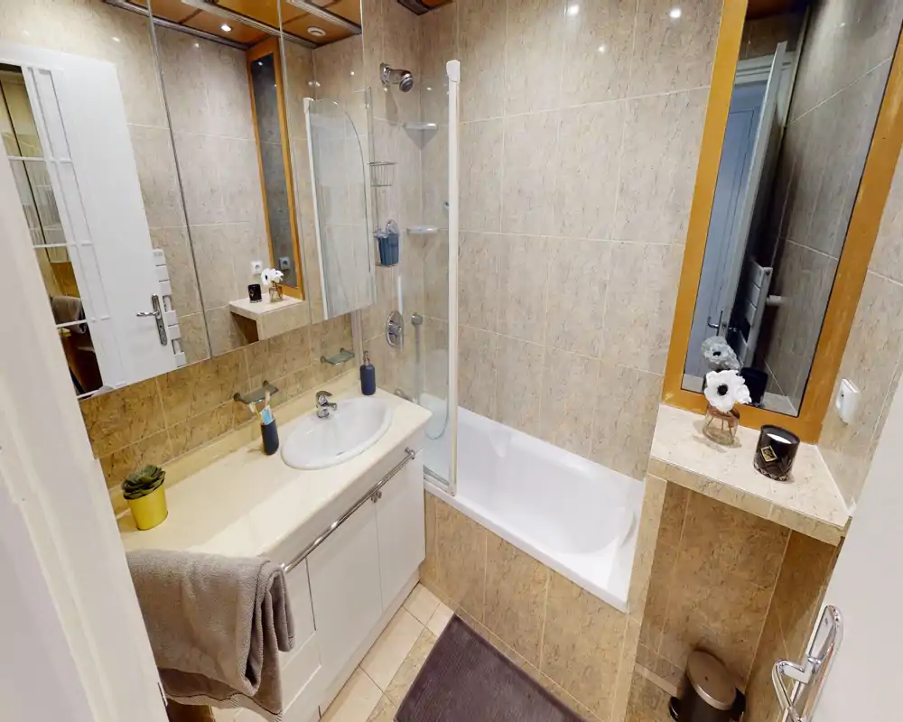 Compact, well-lit bathroom with a bathtub and shower screen, sink with vanity unit, large mirror and neutral tile finishes.
