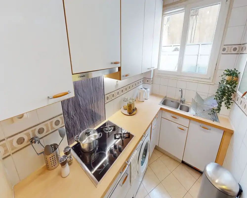 Compact, well-equipped kitchen with a built-in stovetop, sink with draining rack, washing machine under the counter, and bright window lighting.