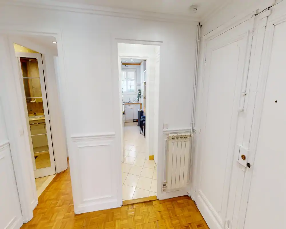 Bright apartment entrance/hallway with white paneled walls, parquet flooring and a view through to the kitchen; clean, neutral decor ideal for showcasing circulation space.