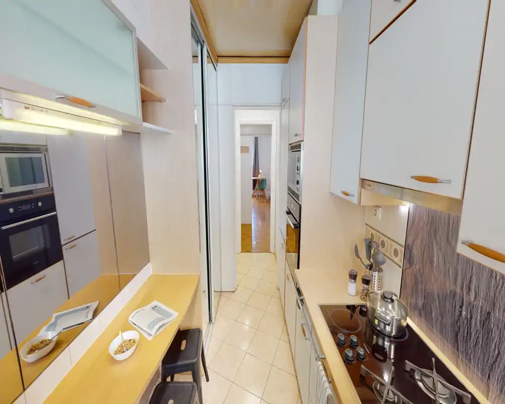 Narrow fitted kitchen with built-in oven, induction hob and a small breakfast counter. Light wood cabinets and tiled floor create a clean, functional layout ideal for compact apartments.