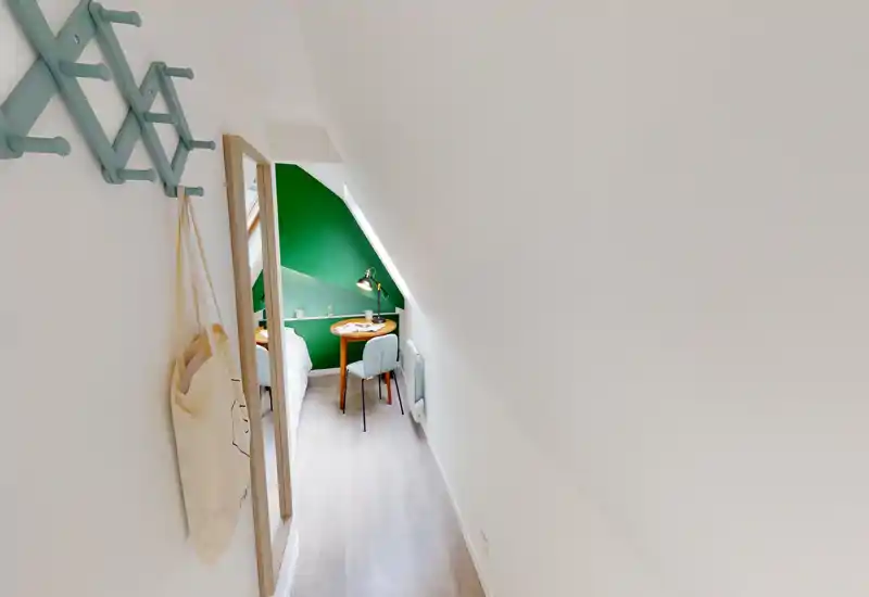 Small attic bedroom seen down a narrow corridor, with a partial view of the bed, green accent wall, a bedside table with lamp and chair — compact and stylish sleeping nook.