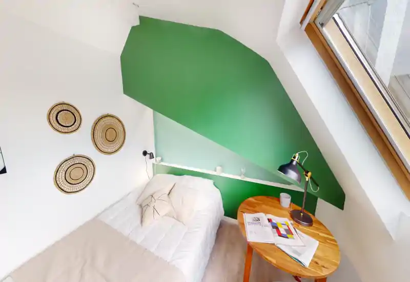 Sunny single bedroom with a neatly made bed under a sloped green-accent wall, wooden side table and large window providing abundant natural light—cozy and well styled for a rental listing.
