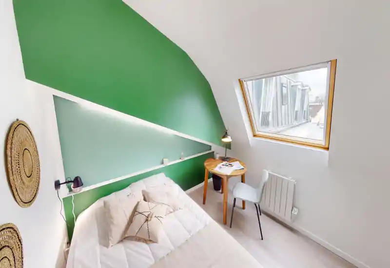 Cozy, well-lit small bedroom with a comfortable double bed, green accent wall and a skylight providing abundant natural light. A compact wooden side table and chair create a neat workspace next to the bed.