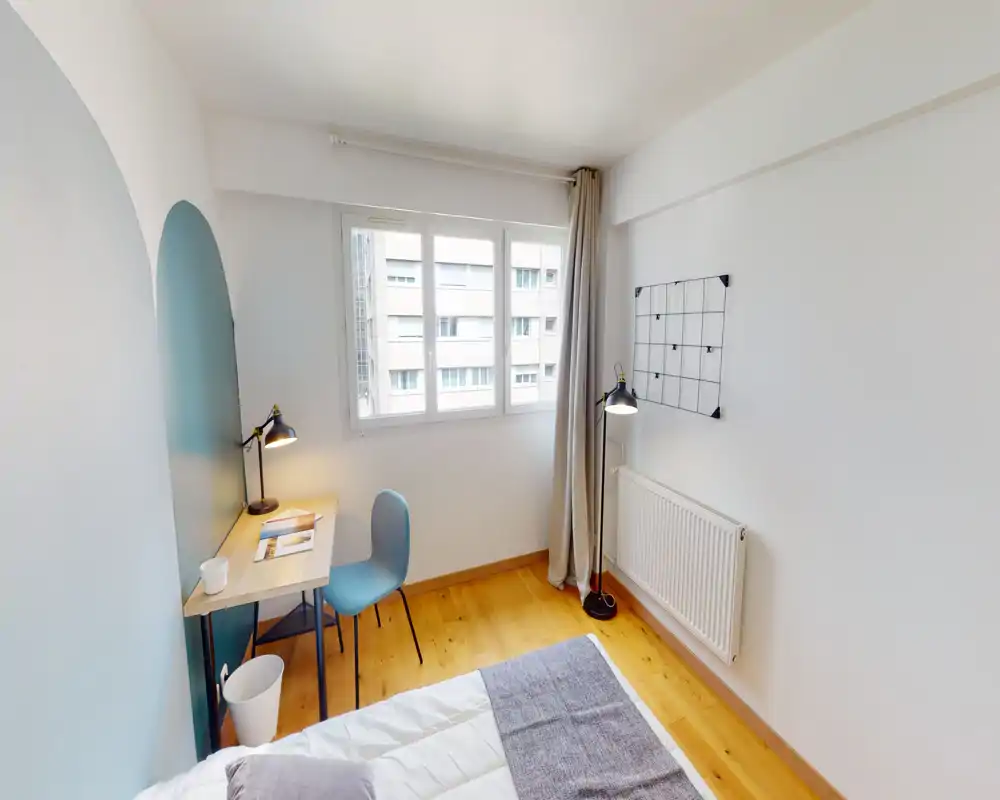 Camera da letto compatta e luminosa con scrivania angolare e vista parziale del letto. La luce naturale dalla finestra, il pavimento in legno caldo e i tessili morbidi creano un ambiente tranquillo e funzionale, ideale per una persona.