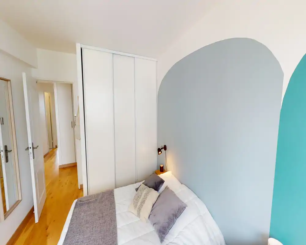 Camera da letto luminosa e moderna con letto matrimoniale ben rifatto, cuscini decorativi e una lampada da comodino. Parete dipinta dai toni pastello e pavimento in legno creano un ambiente caldo e di stile.