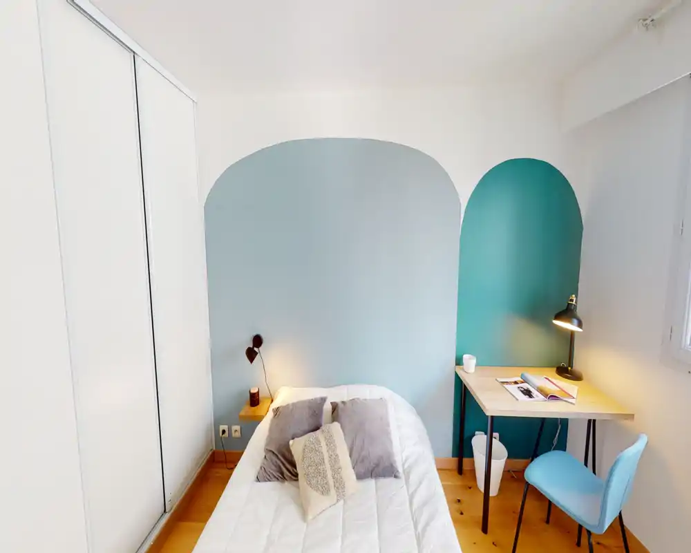 Camera da letto moderna e luminosa con letto singolo ben rifatto, cuscini decorativi e una piccola scrivania vicino alla finestra. La parete con archi nei toni turchese e azzurro crea un'atmosfera elegante e accogliente.
