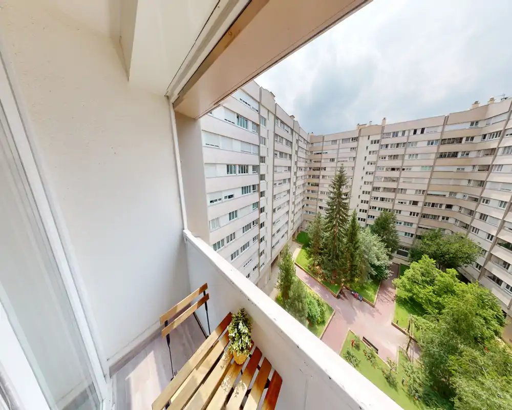 Balcon privé ensoleillé donnant sur une cour paysagée avec arbres et allées ; petite table en bois et plante en pot offrent un coin agréable pour se détendre.