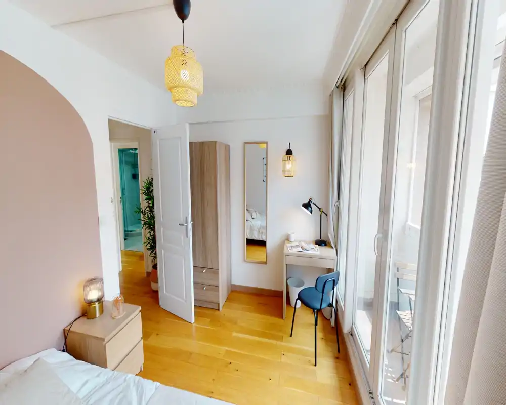 Chambre moderne et lumineuse avec parquet, table de chevet et lampe, armoire et un petit bureau près de grandes fenêtres donnant sur le balcon — ambiance chaleureuse et accueillante, idéale pour une annonce locative.