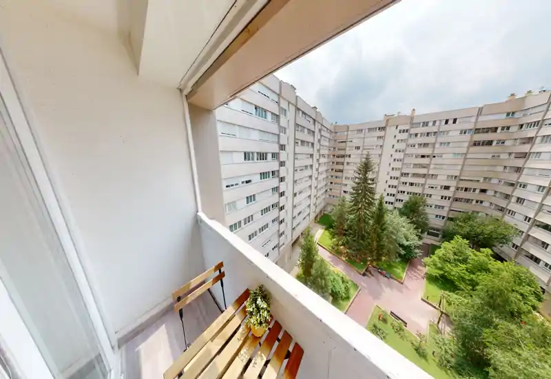 Sunny private balcony overlooking a landscaped courtyard with mature trees and pathways; small wooden seating and potted plant create a pleasant spot to relax.