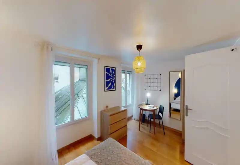 Bright, cozy bedroom with wooden floors, a visible bed in the foreground, two large windows and a small desk area — ideal for single or couple stays.